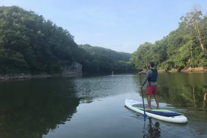 Person paddleboarding on a calm river surrounded by green hills and trees.