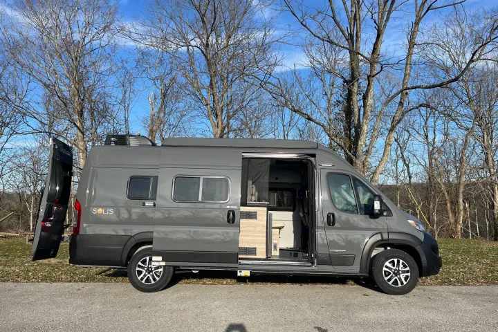 Gray campervan with open sliding door, parked on paved area near leafless trees.